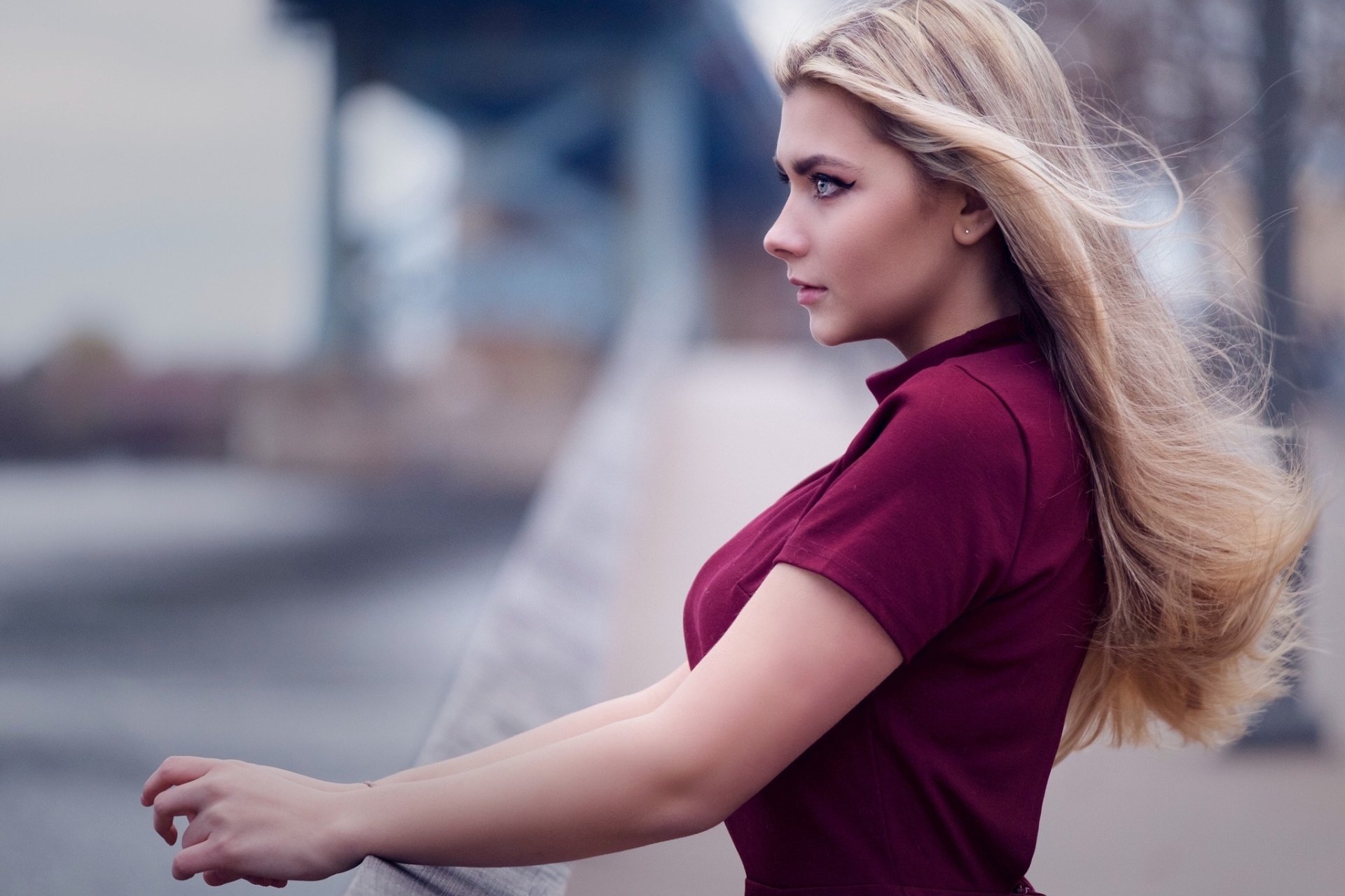 A woman model stands gracefully by a railing, her long hair flowing in the wind, gazing thoughtfully into the distance with a blurred background of urban scenery.