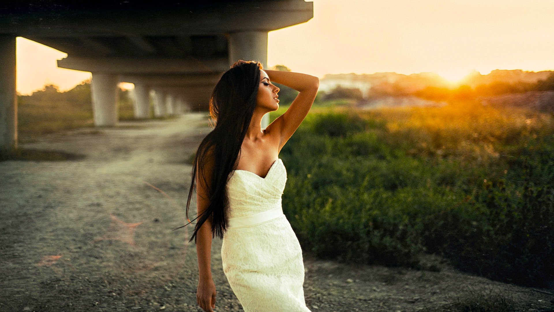 A woman in a white dress stands gracefully under a bridge, basking in the summer sun. Her long hair flows as she poses against a backdrop of greenery and a glowing sunset.