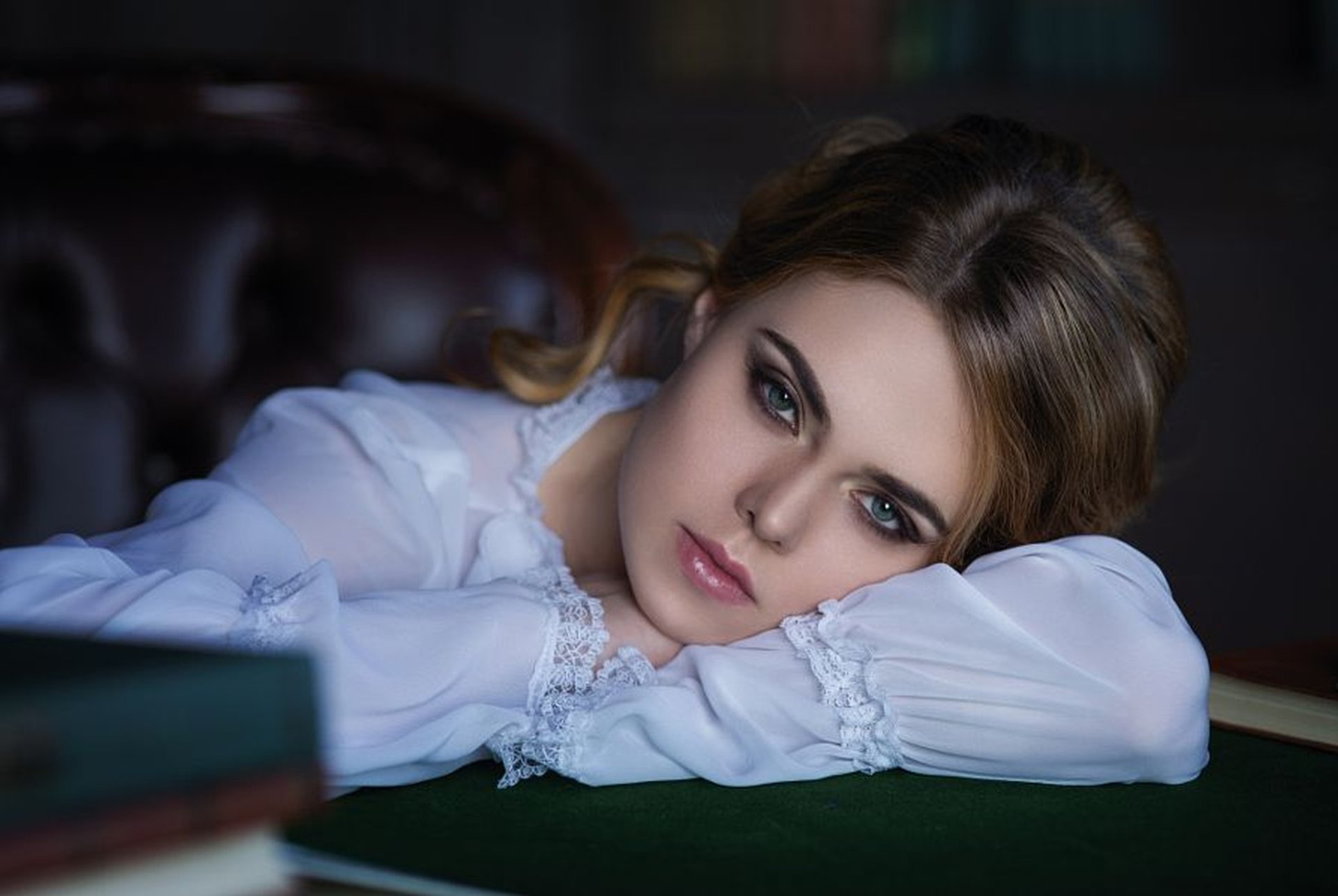 A woman model gazes thoughtfully while resting her arms on a table, showcasing her elegant attire and striking makeup against a softly blurred background.