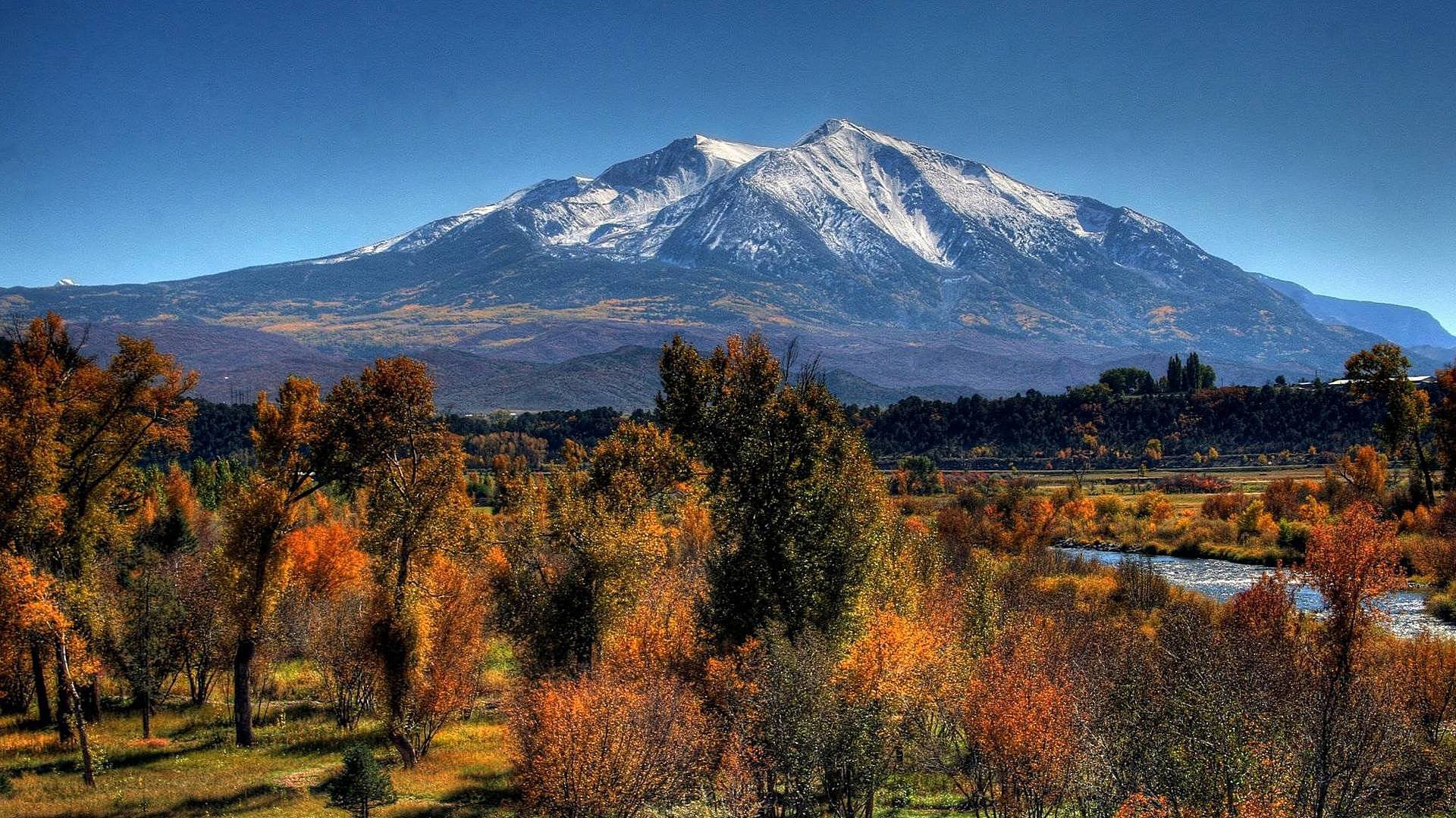 Snow-capped mountain rises behind a colorful autumn landscape of trees and a winding river under a clear blue sky, showcasing vivid nature scenery.