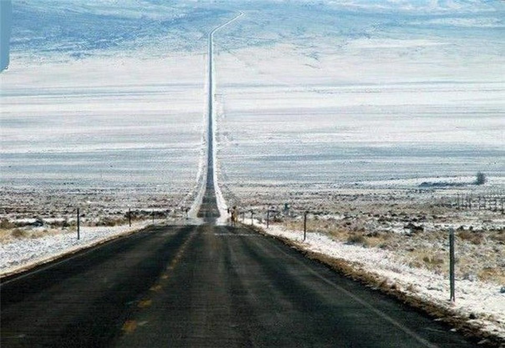 A long, straight road stretches into a snowy landscape, showcasing a stark contrast between the man-made path and the vast, empty terrain surrounding it.