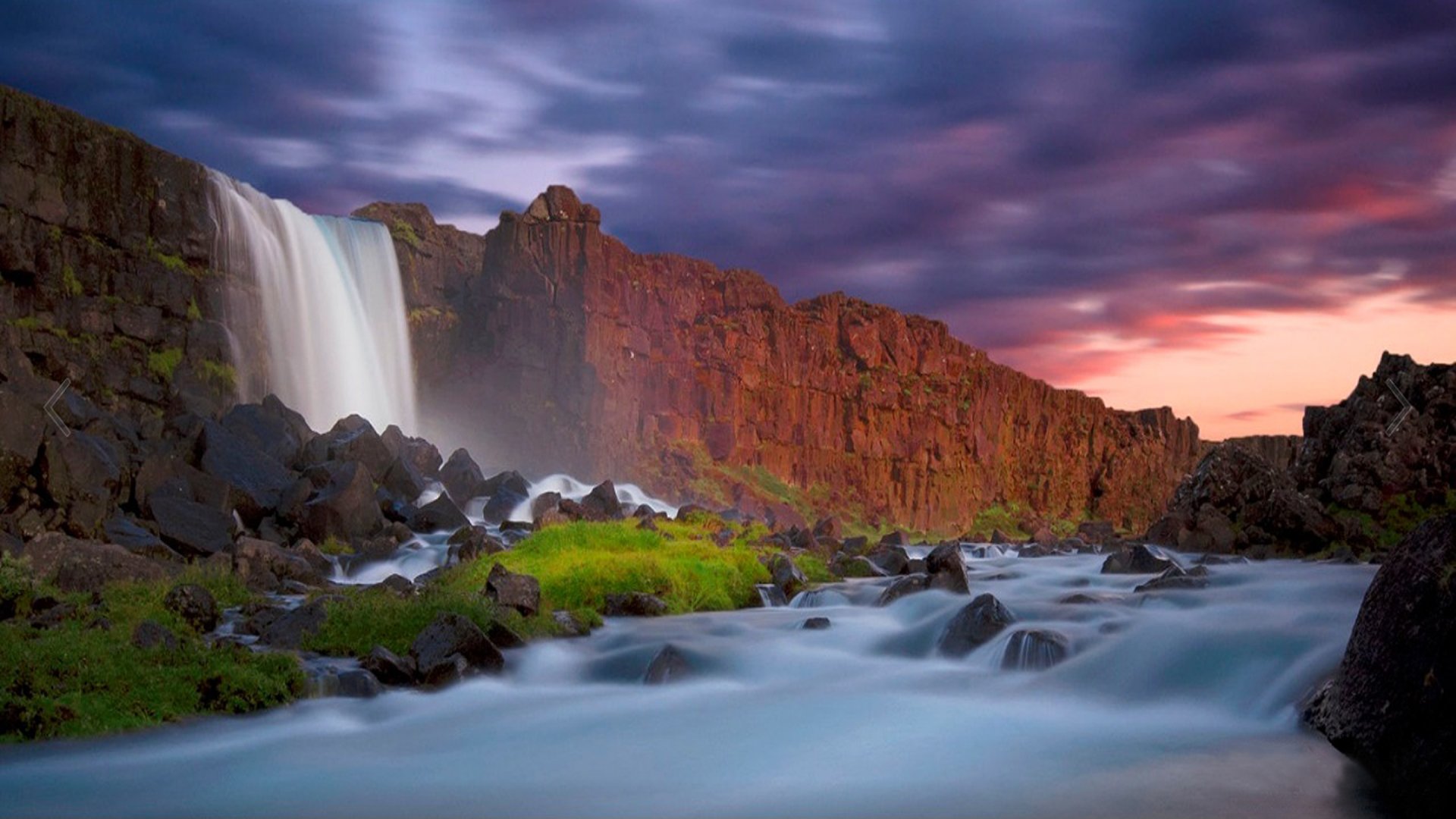 A waterfall cascades down a rocky cliff with lush greenery by the water’s edge under a colorful sky at sunset.