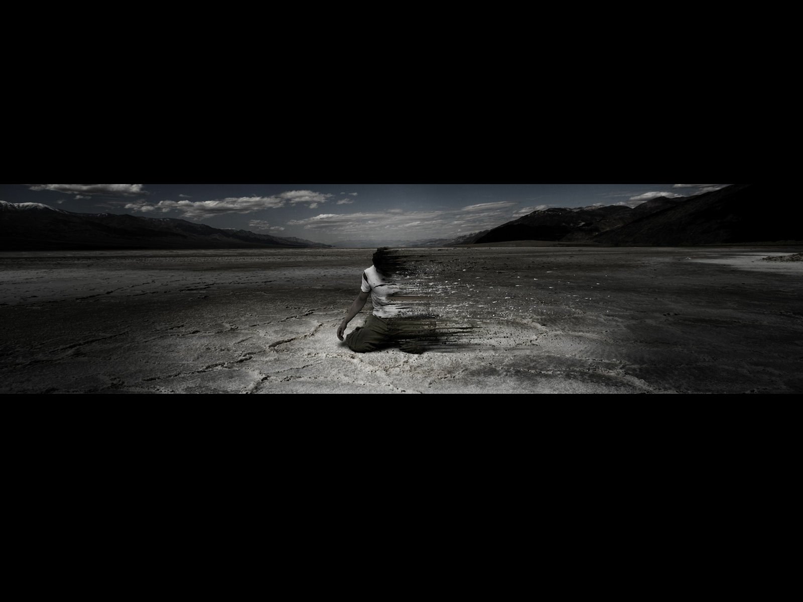Artistic black-and-white image of a solitary human figure seated on cracked ground in a vast, desolate landscape under a cloudy sky.