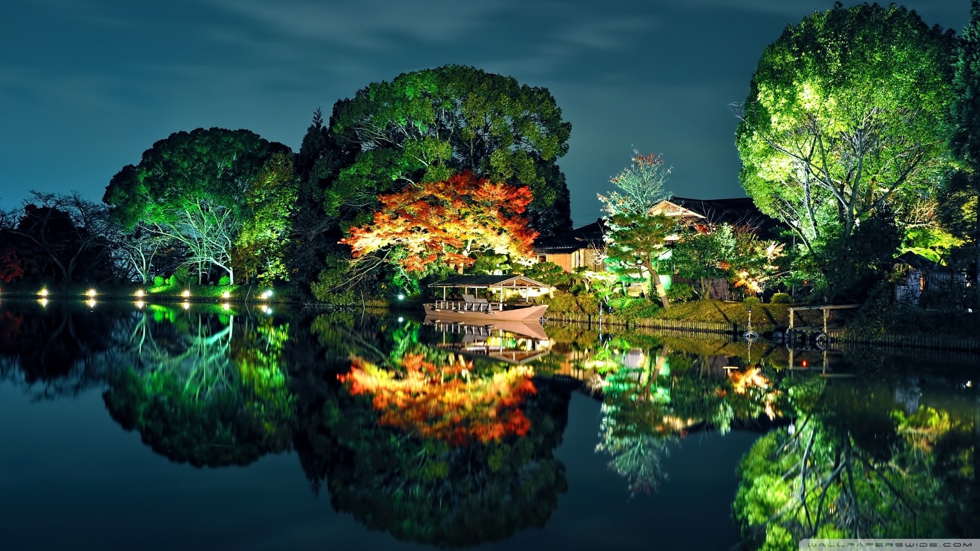 Night photography of illuminated lakeside trees and a pavilion, their colors mirrored in a glassy reflection on the water.