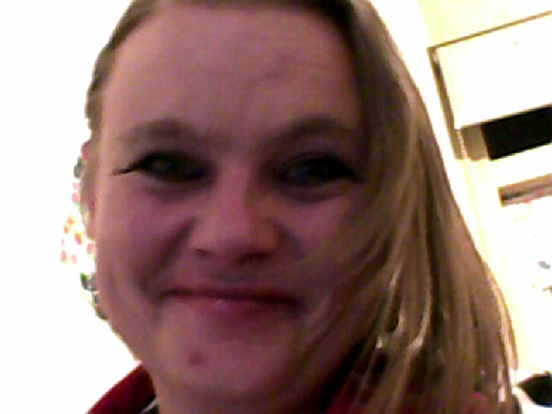 Close-up of a smiling woman's face with light hair swept to one side and dark eyeliner, indoors near a window.