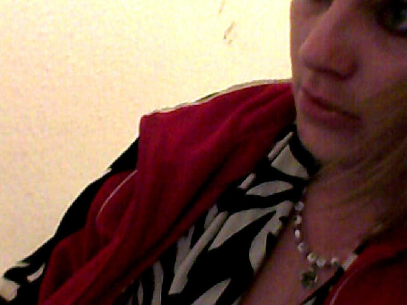 Close-up of a woman's face and upper torso in a red jacket, patterned top and necklace, gazing to the right against a pale wall.