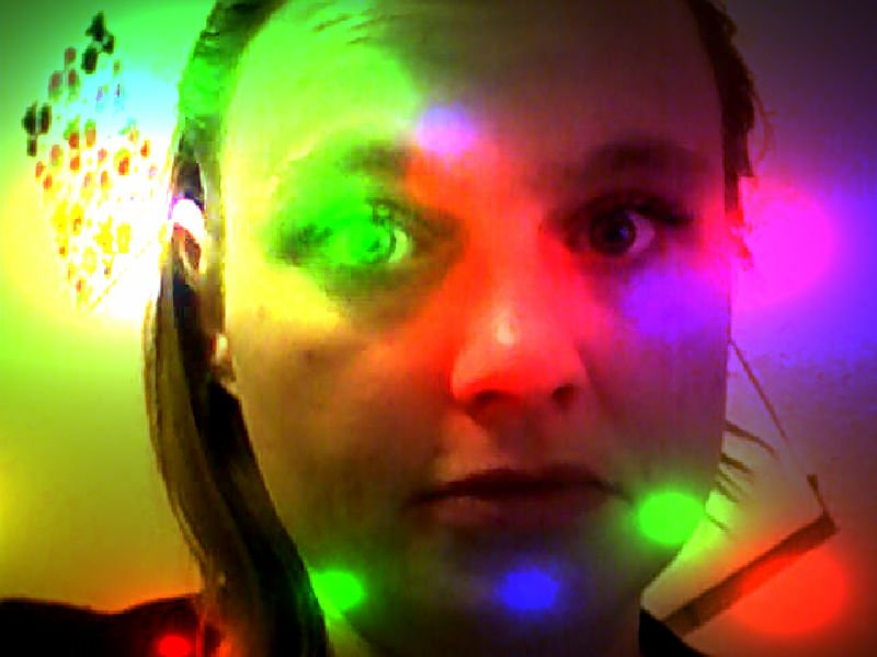 Close-up of a woman's face lit by multicolored green, red and purple lights, with bright spots and reflections across her cheeks and forehead.
