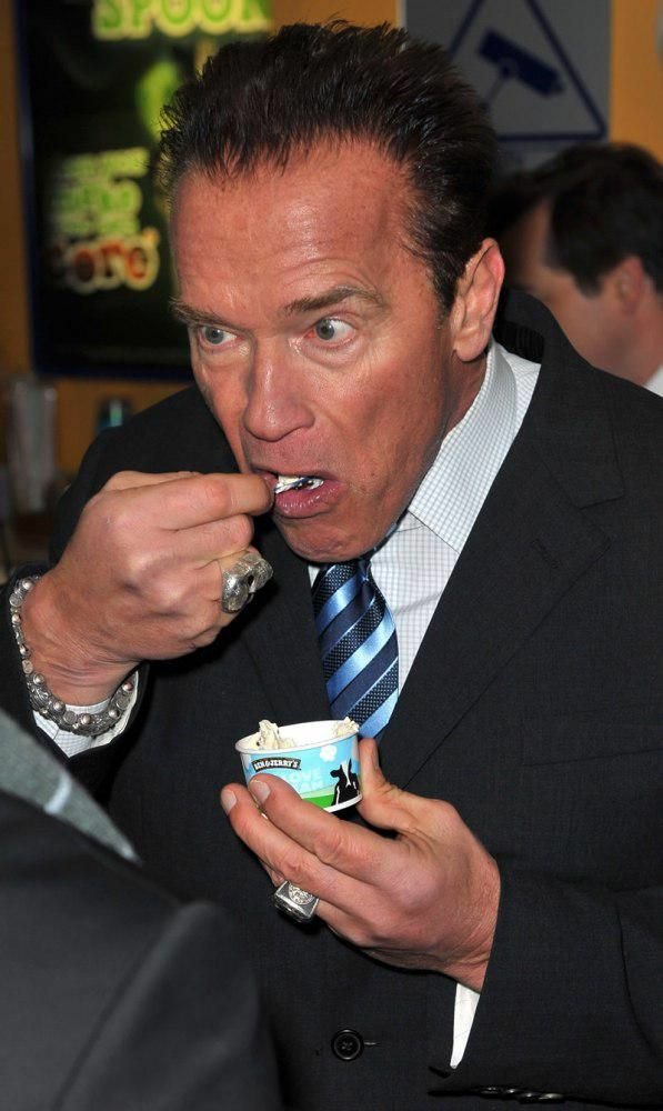 Arnold Schwarzenegger enjoys ice cream at an event, eagerly taking a bite while dressed in a suit, showcasing a candid moment of indulgence.