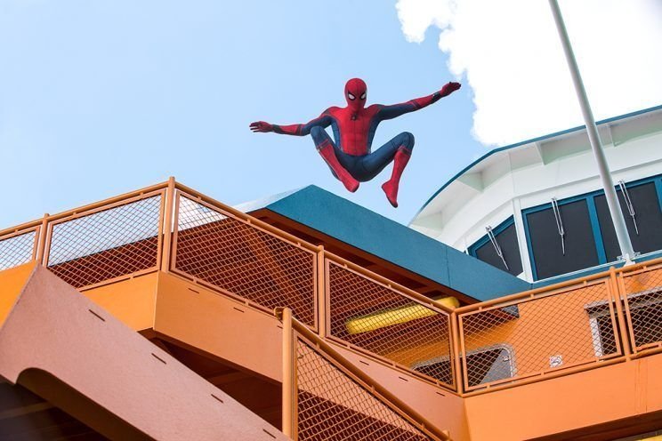 Spider-Man in his iconic suit leaps from an orange staircase structure against a clear blue sky, capturing a dynamic moment from the movie Spider-Man: Homecoming.