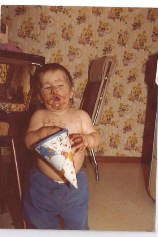 Vintage photograph of a young child with a party hat, face and hands covered in cake, standing in a room with patterned wallpaper.
