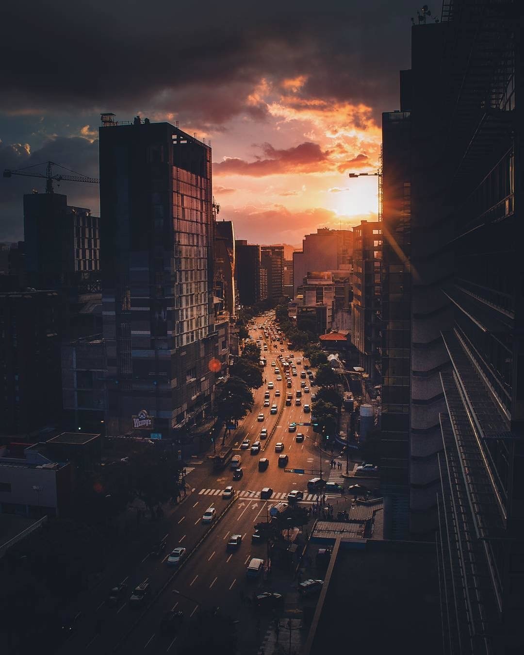 A man-made cityscape at sunset with tall buildings lining a busy street filled with cars and illuminated by the warm glow of the setting sun.