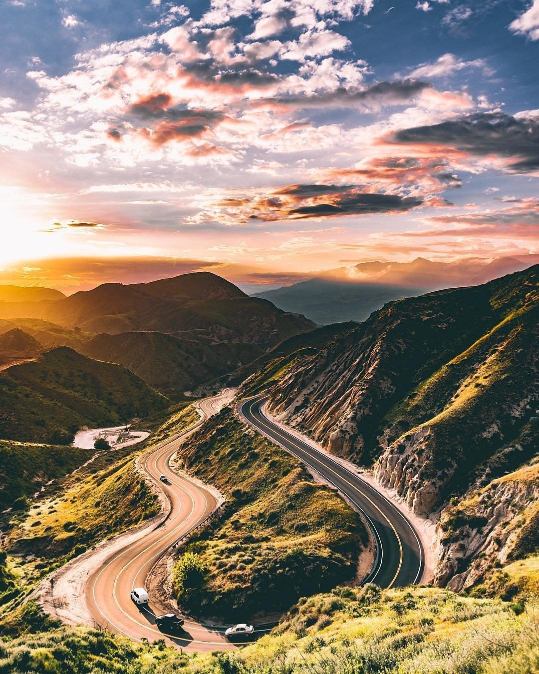 Winding man-made road snakes through green mountainous landscape under a colorful sunset sky with scattered clouds.