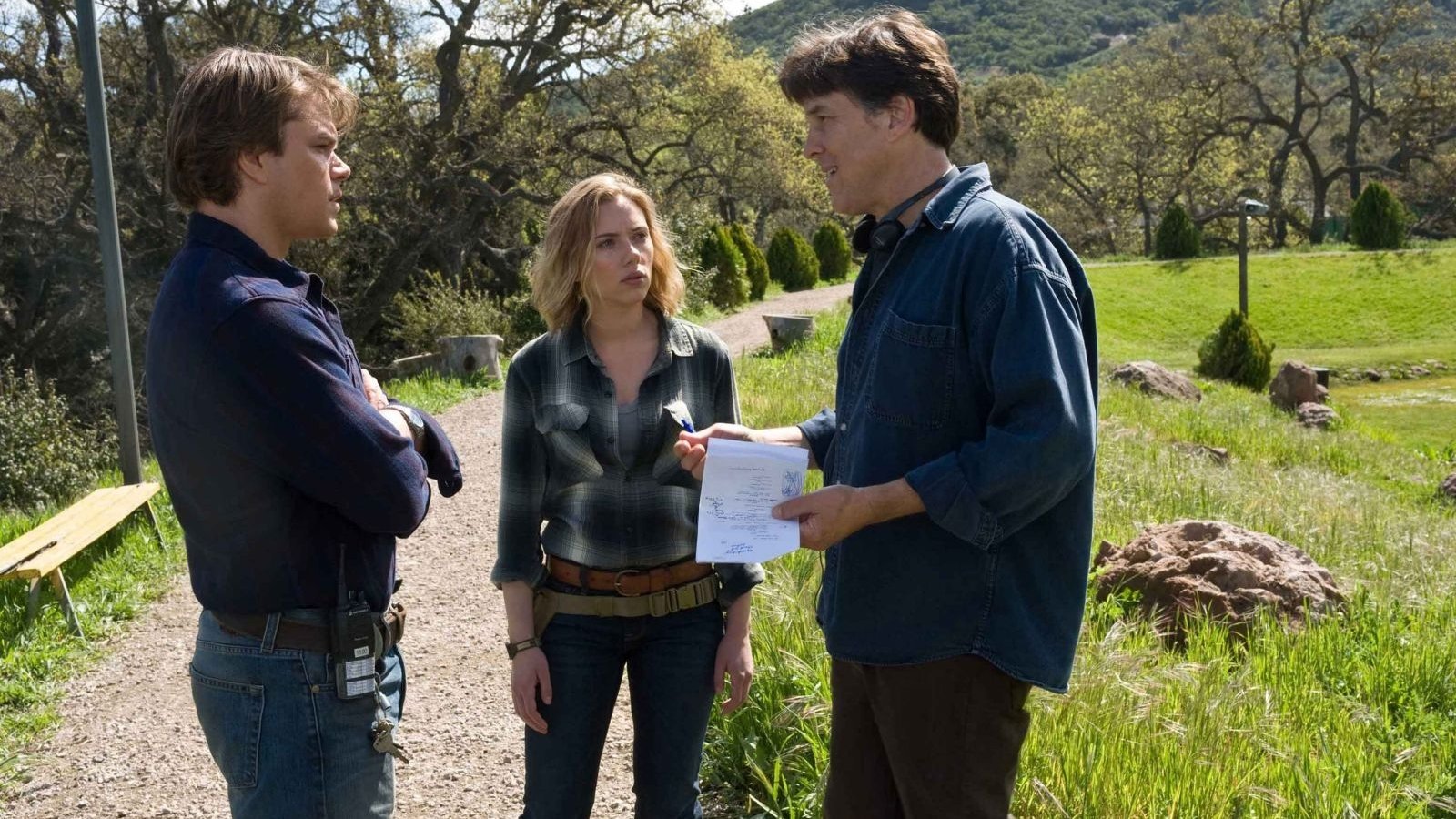 Scene from the film We Bought a Zoo: three characters on a grassy path outdoors — two men and a woman converse while one man holds a sheet of paper.