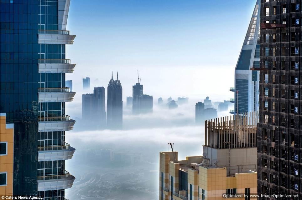 Fog envelops a man-made cityscape with towering skyscrapers rising above the mist under a clear blue sky.