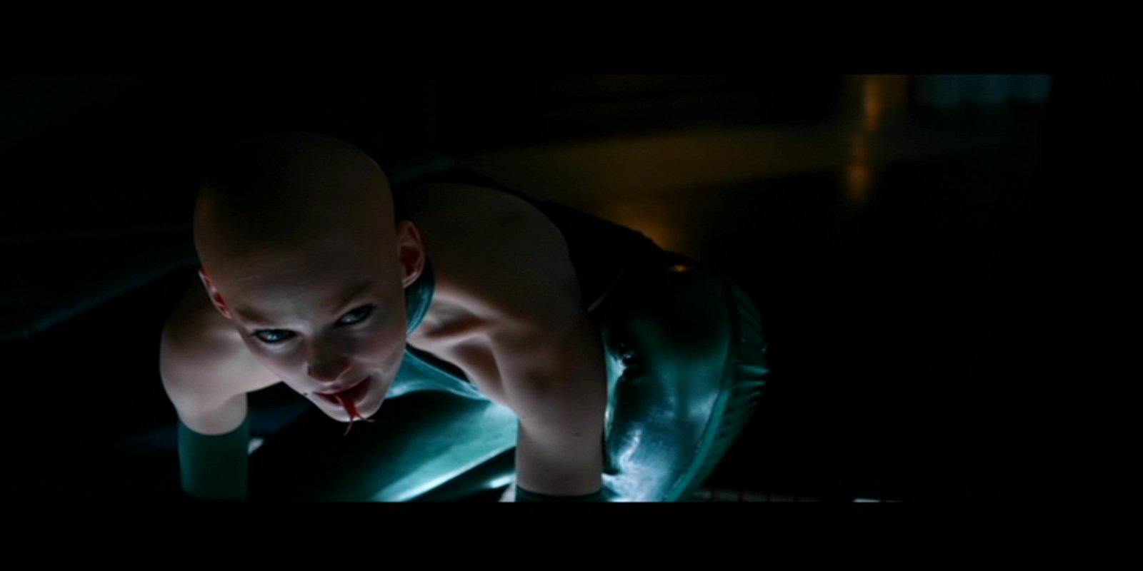 A character from the movie The Wolverine is depicted crawling in a darkened space, showcasing a bald head and a striking teal outfit, creating a sense of tension and intrigue.