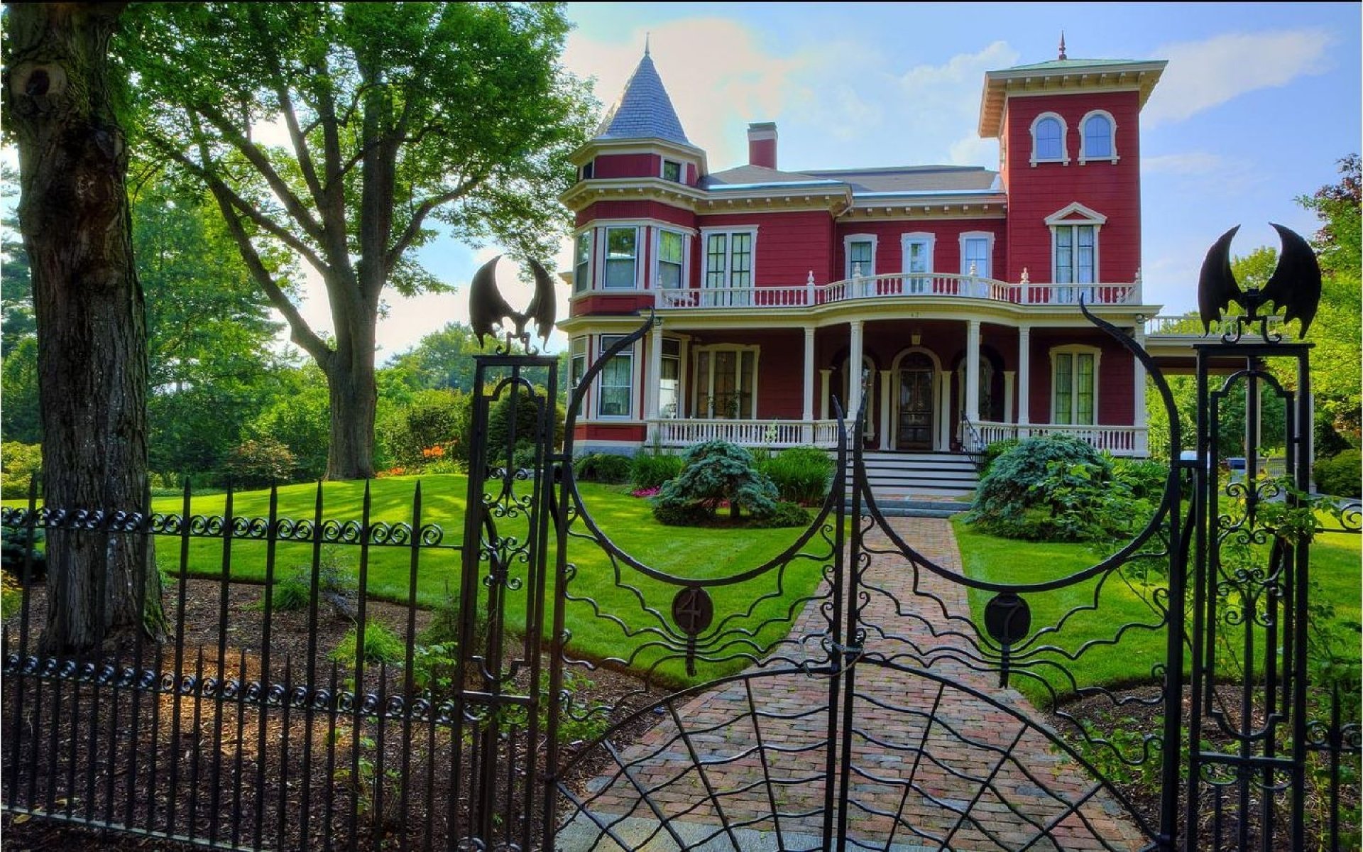 A grand Victorian house with intricate architecture stands behind a decorative metal gate, surrounded by lush greenery and trees. The vibrant red exterior adds charm to the scene.