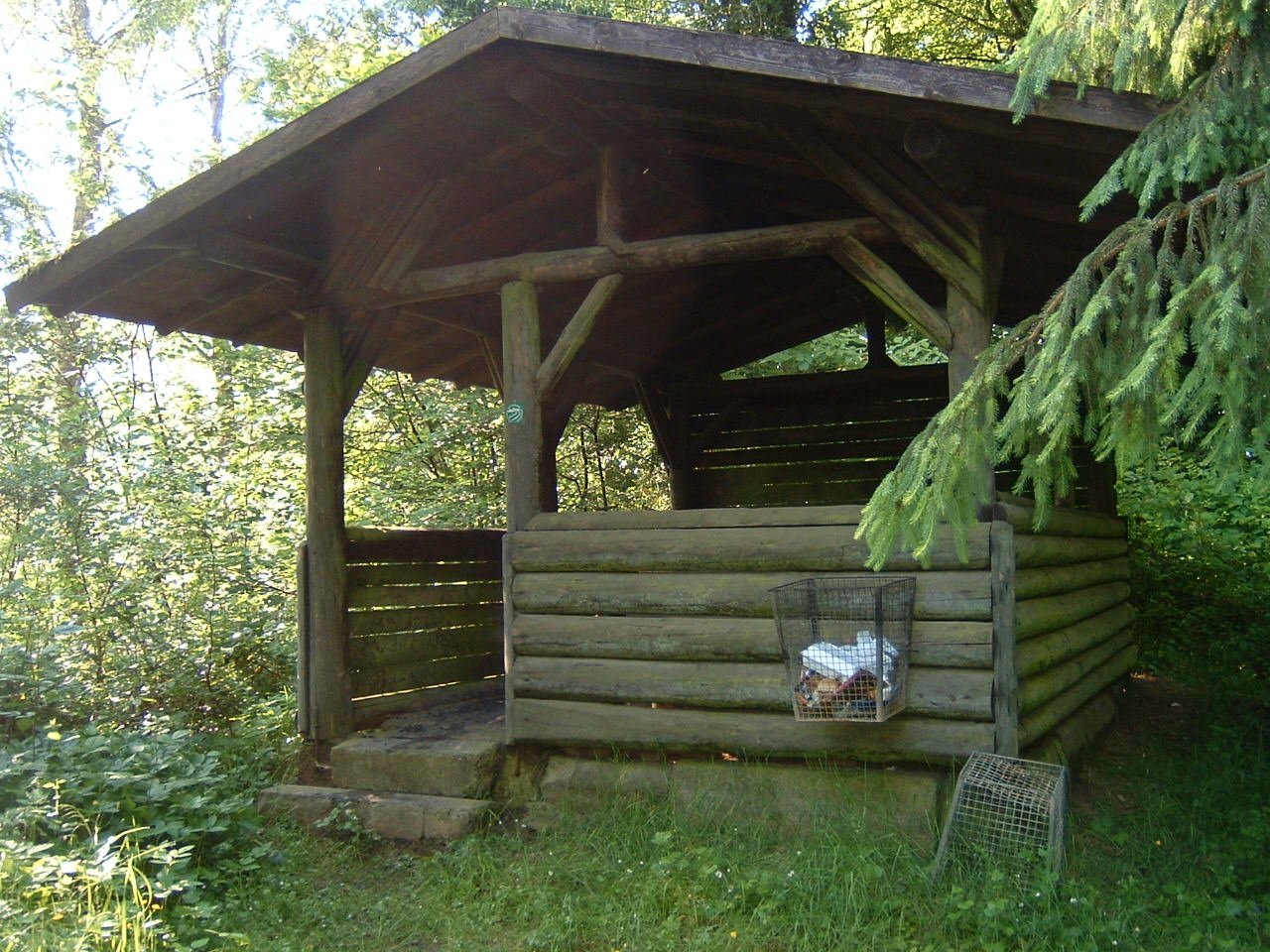 A man-made rustic log cabin shelter with open sides, sturdy wooden posts and a pitched roof, set on a grassy edge of a wooded area.