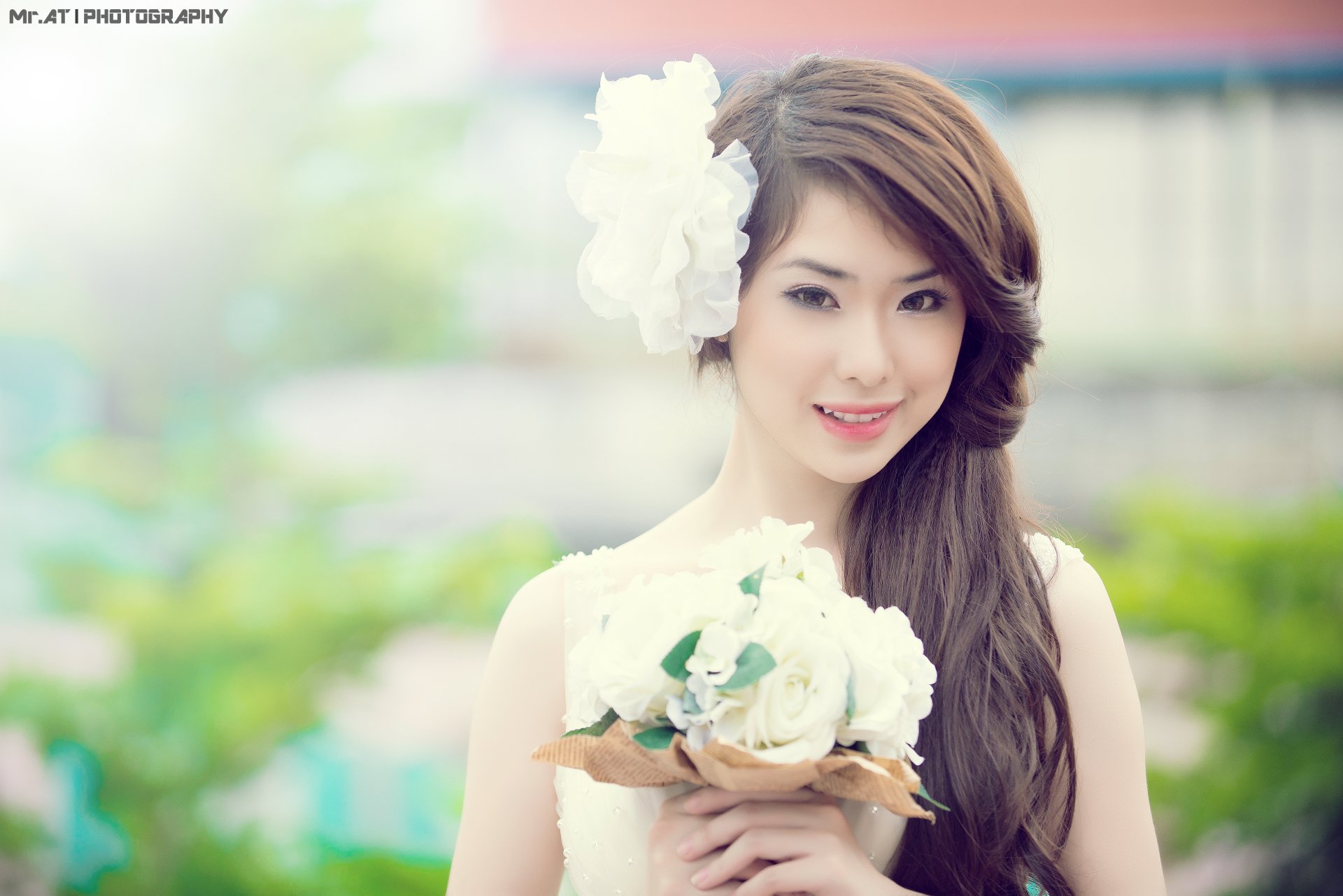 Portrait of an Asian woman with long hair adorned with a white flower, holding a bouquet of white flowers, against a soft, blurred outdoor background.