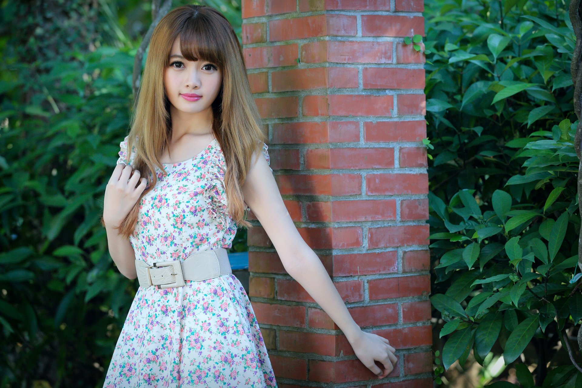 Young woman with long brown hair in a white floral sundress and wide belt, leaning against a brick pillar surrounded by green foliage.