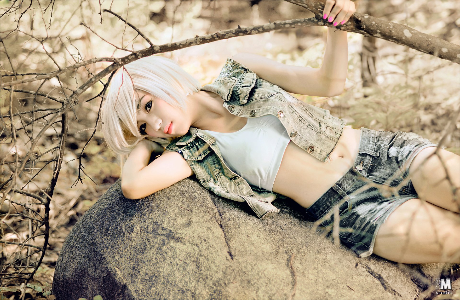 Young woman with short platinum-blond hair reclining on a rock in a sunlit wood, wearing a cropped white tank, denim vest and shorts.