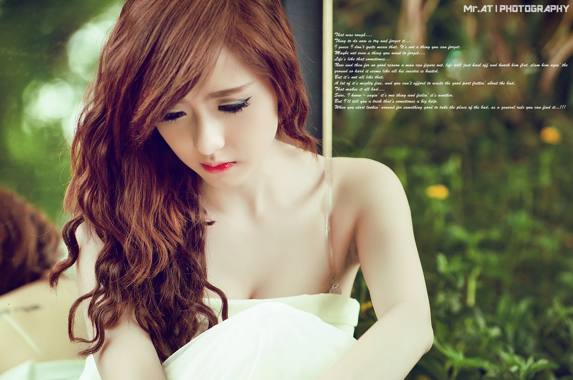 Asian woman with long wavy brown hair in a white dress sitting outdoors among green grass, gazing downward with a contemplative expression.