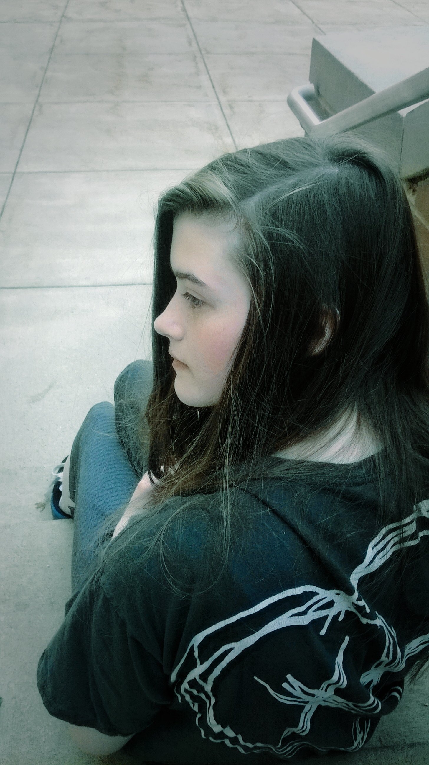 Abstract, beautiful portrait of a seated person in profile on concrete steps, long dark hair over a graphic black shirt, soft muted tones and a minimalist, contemplative composition.