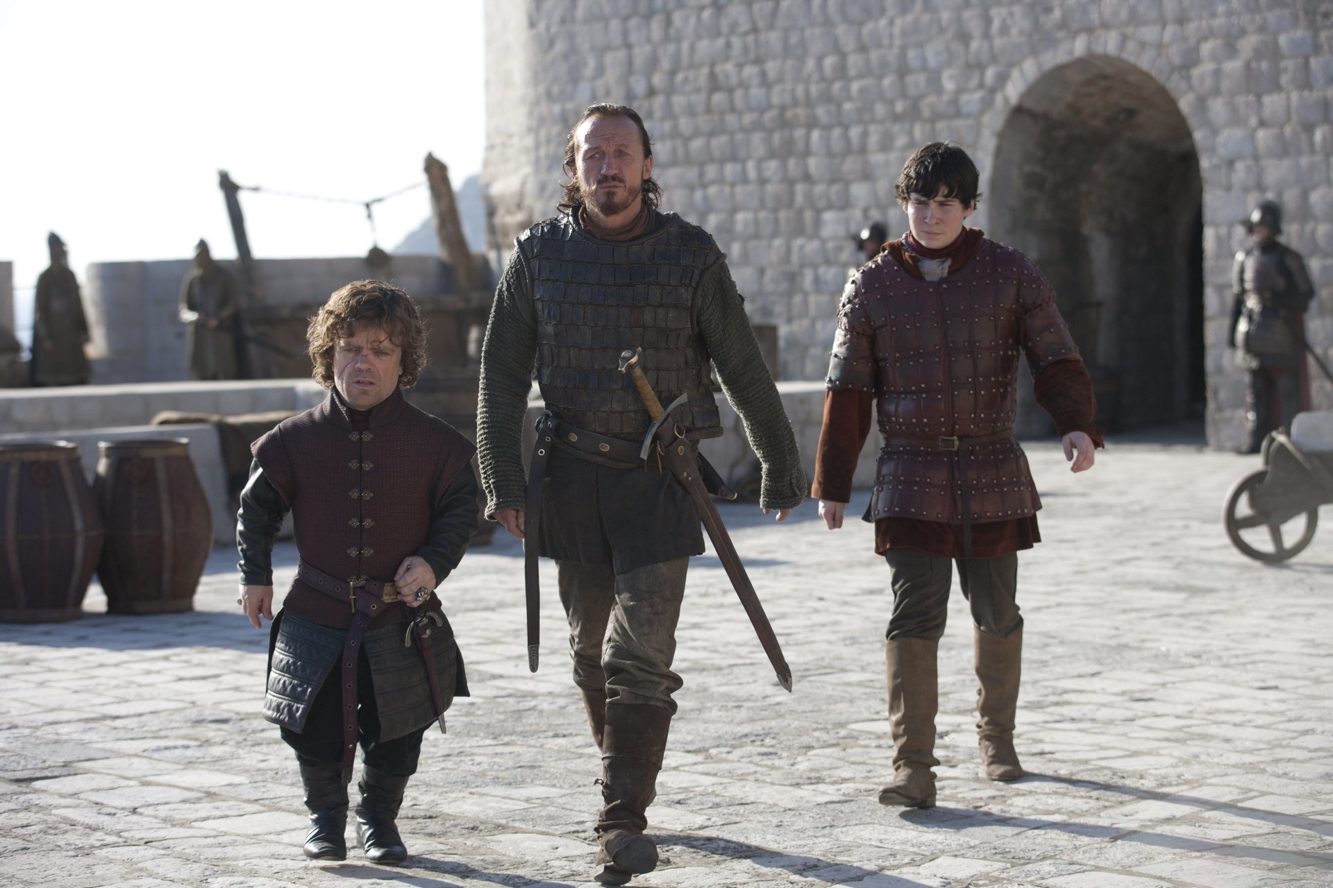 Peter Dinklage as Tyrion Lannister, Jerome Flynn as Bronn, and Daniel Portman as Podrick Payne walking together in a courtyard in the TV show Game of Thrones.