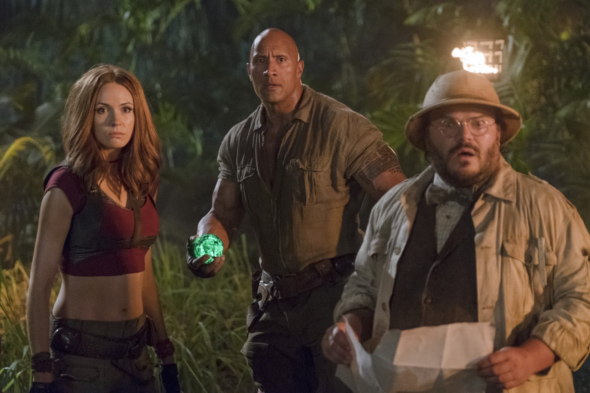 A scene from Jumanji: Welcome to the Jungle, featuring three characters in a lush jungle setting, with one holding a glowing green gem. Adventure and excitement are palpable.