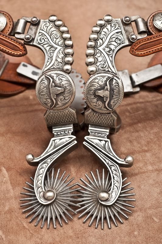 Close-up photography of ornate silver cowboy spurs with engraved floral and steer-head motifs resting on a suede leather backdrop.