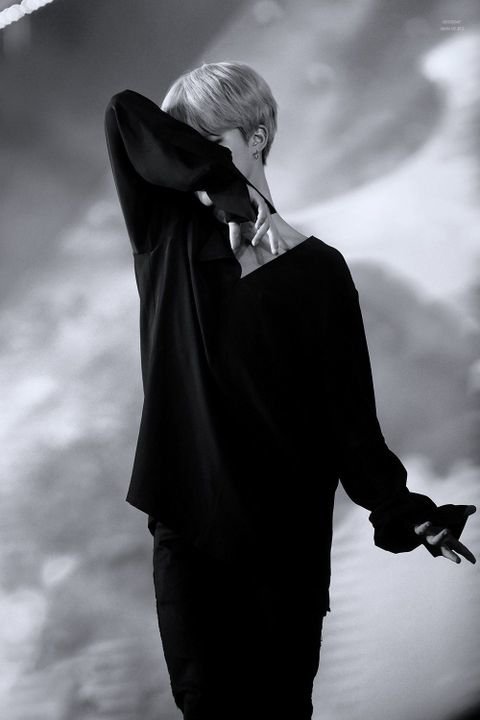 Black-and-white photo of a BTS member performing on stage, partially covering their face with one arm against a blurred background.