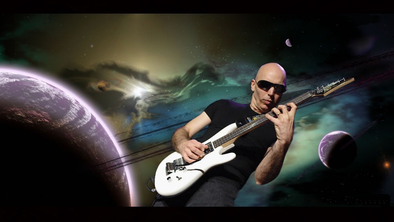 Joe Satriani, wearing sunglasses, performs on a guitar against a cosmic background featuring planets and stars, showcasing his musical talent in a vibrant, otherworldly setting.