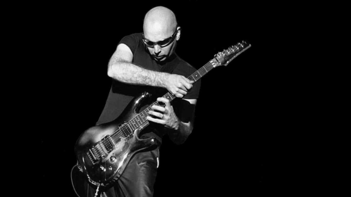 A black and white image of Joe Satriani passionately playing electric guitar, showcasing his dynamic stage presence and musical skill.