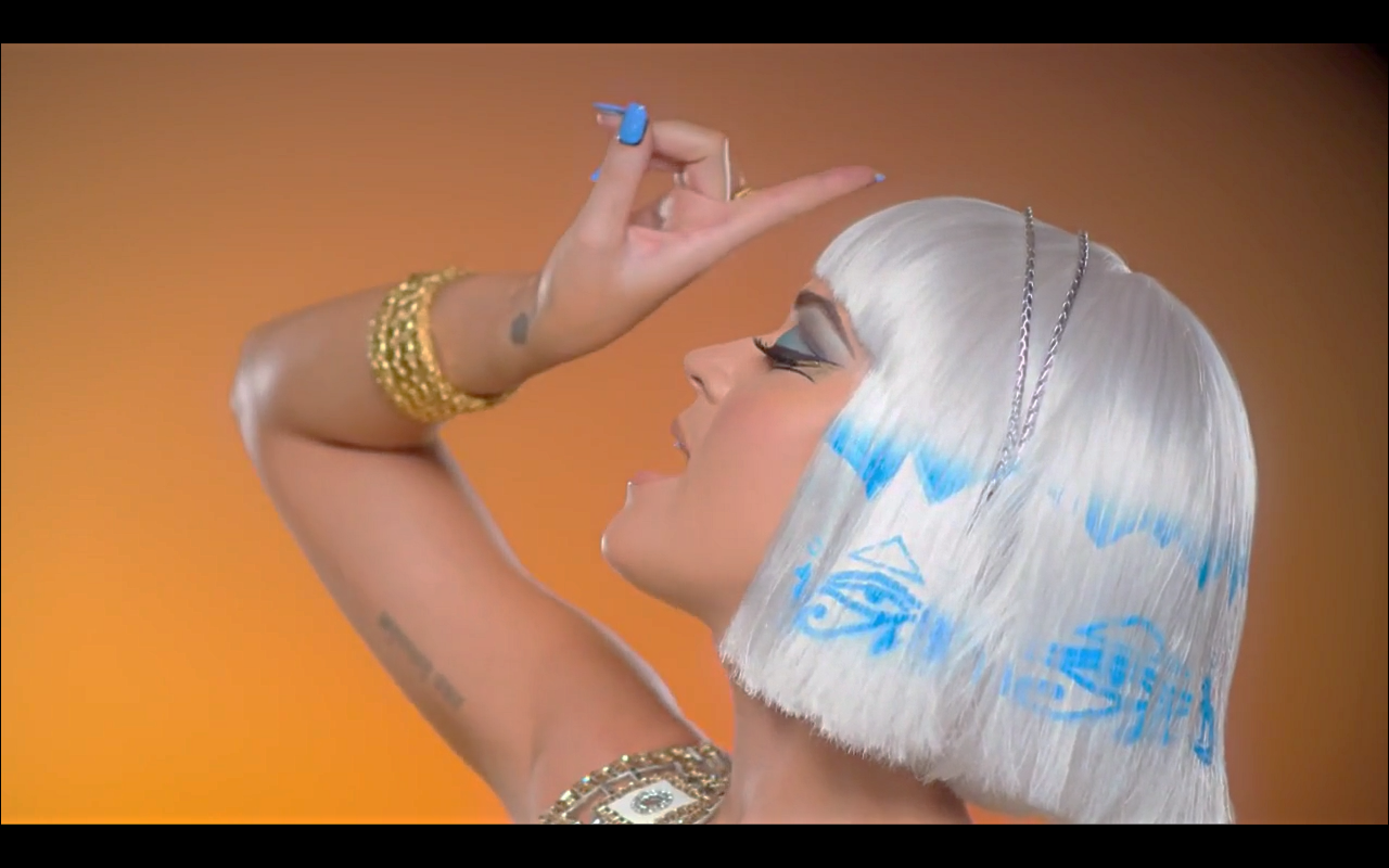 Katy Perry poses gracefully with a striking short white hairstyle, showcasing artistic makeup and accessories against a vibrant orange background, embodying her dynamic music style.