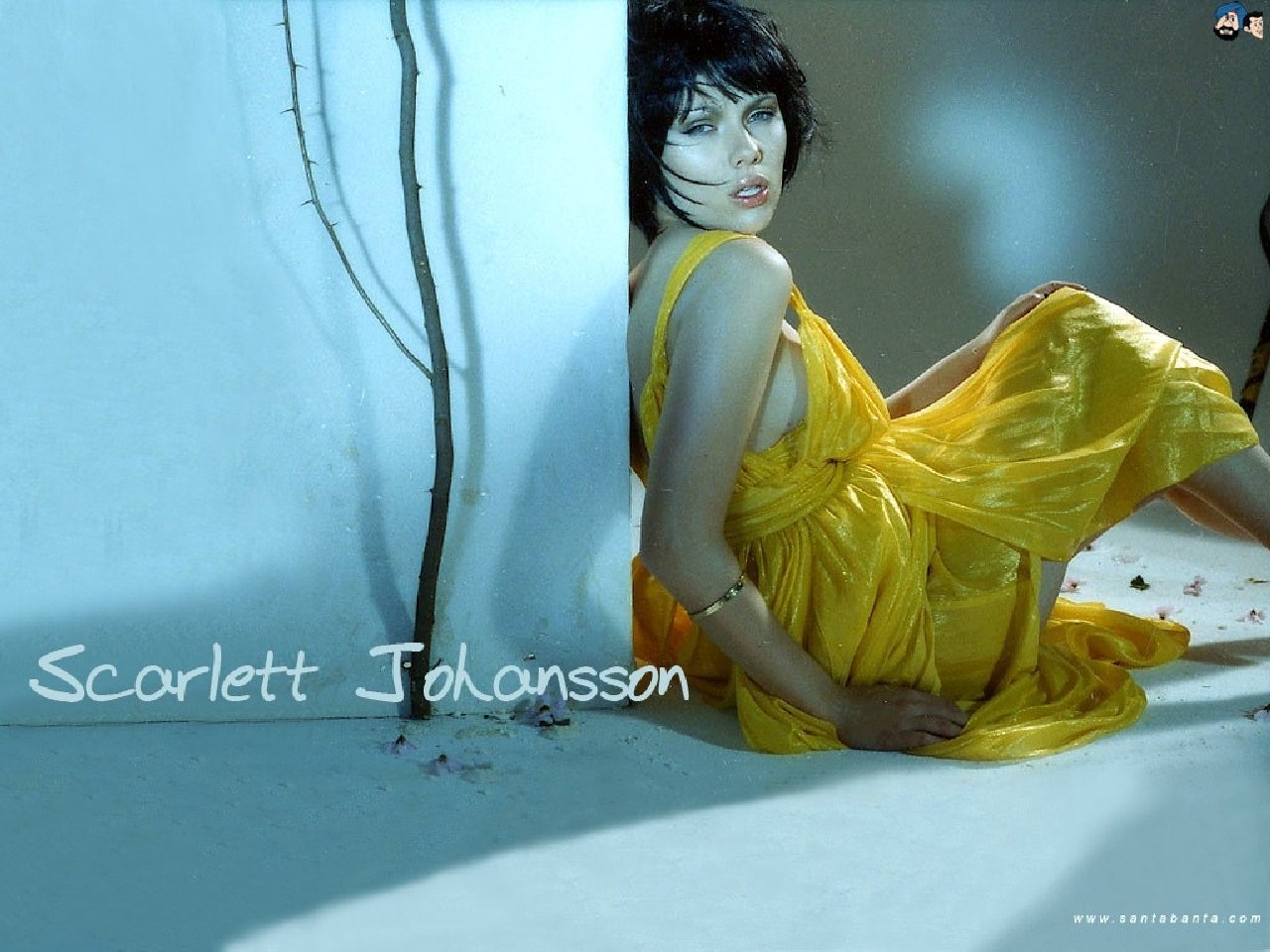 Scarlett Johansson poses gracefully in a striking yellow dress, leaning against a wall, her expression captivating against a softly lit background.