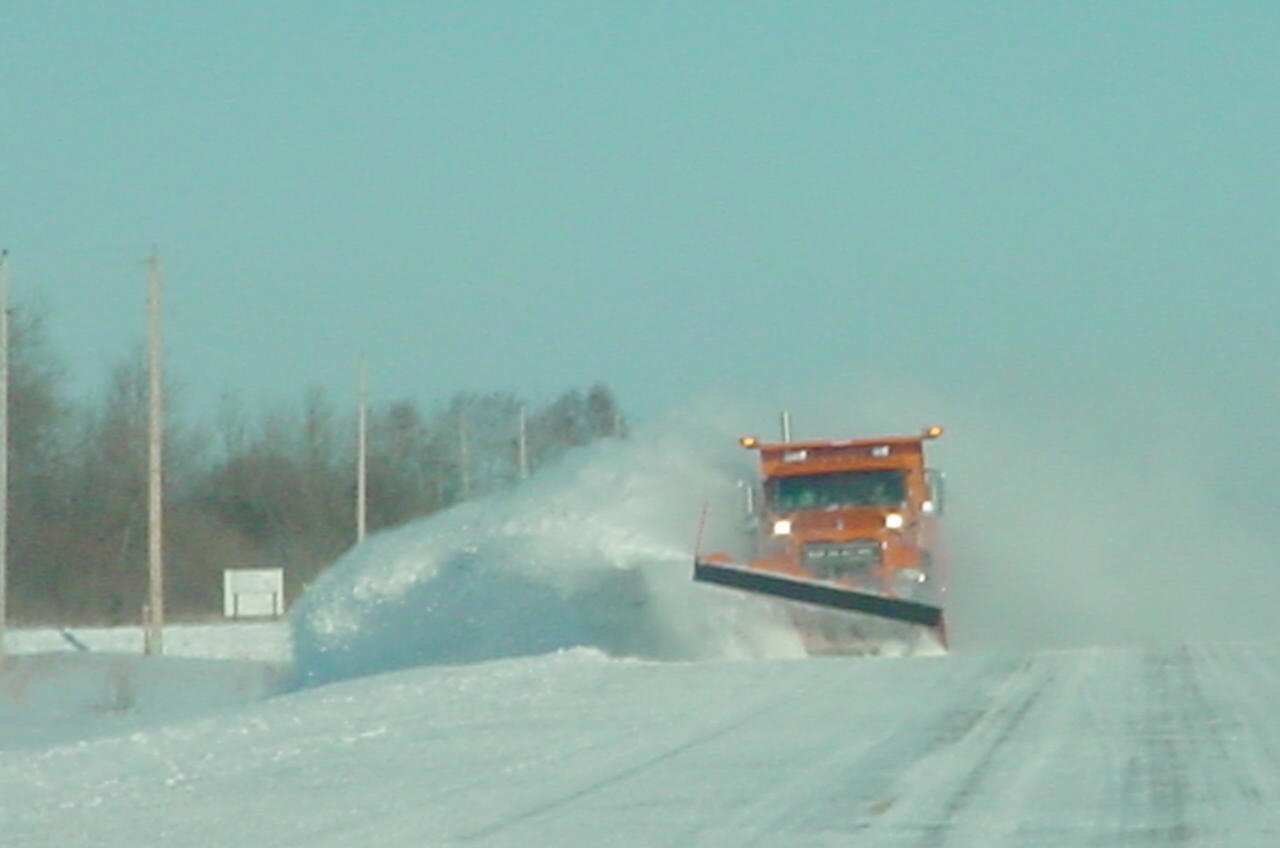 A snowplow vehicle clears snow from a road on a bright, clear day.