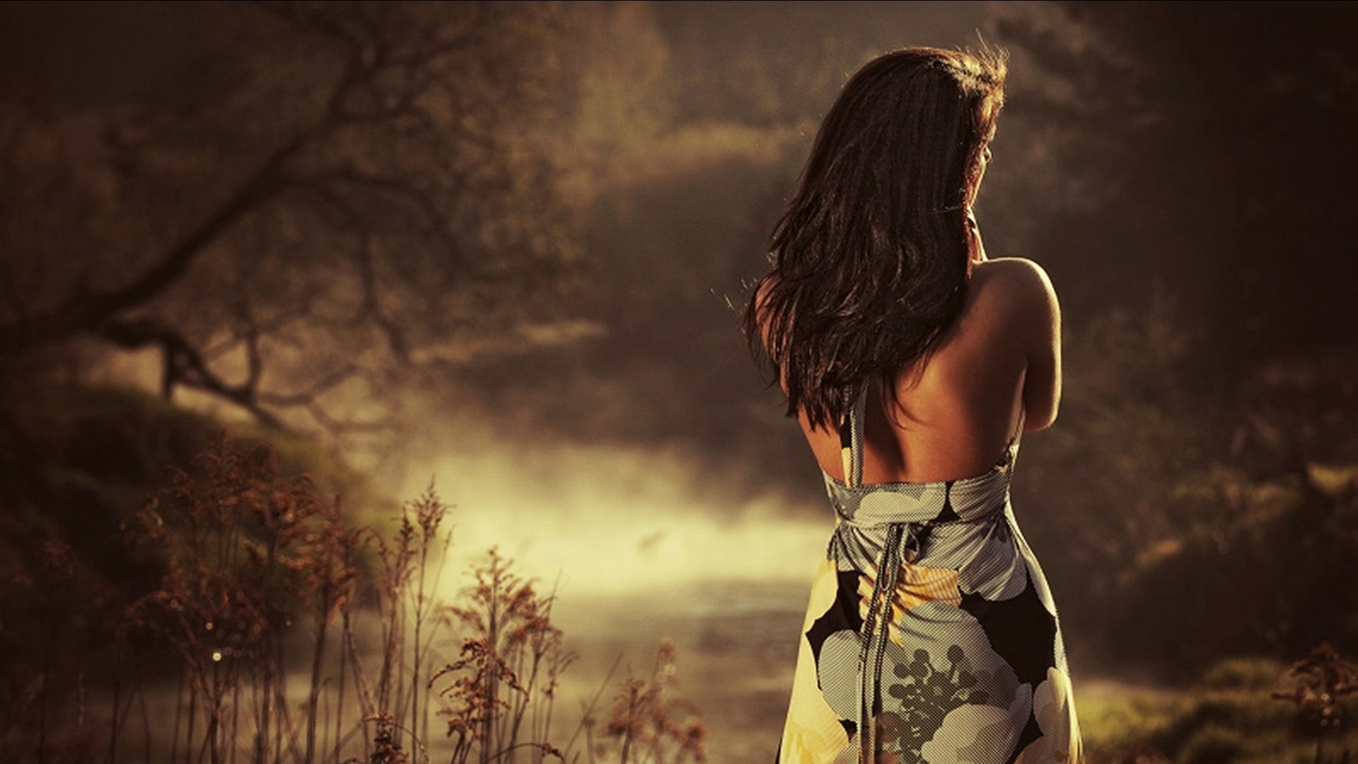 A woman stands with her back to the camera, gazing at a misty landscape, wearing a floral dress that complements the serene atmosphere around her.