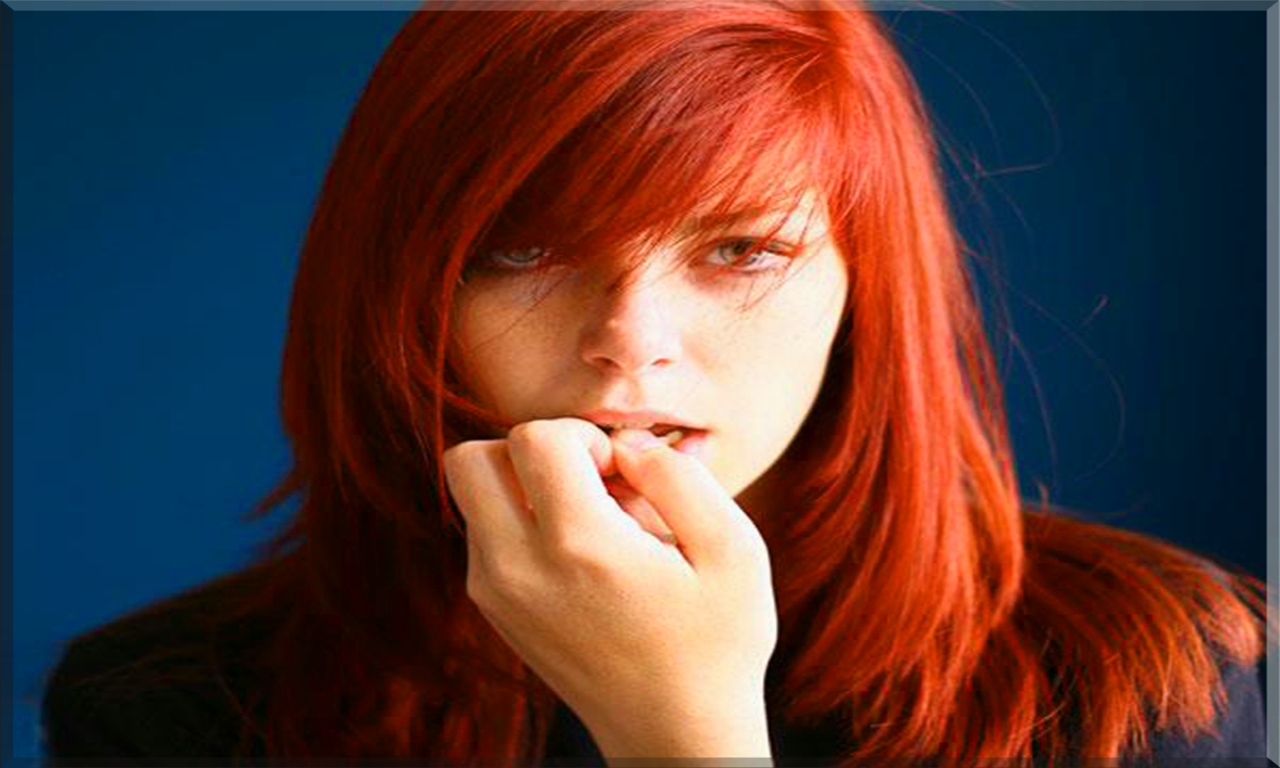 Close-up of a redhead woman model with striking red hair against a deep blue background, resting her hand near her mouth.