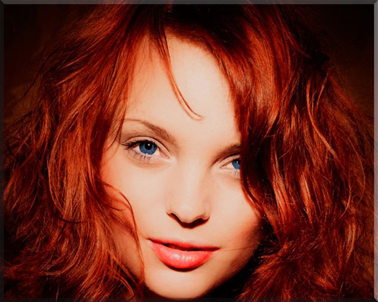 A close-up portrait of Izabella Miko, a redhead model with vibrant curls and striking blue eyes, exuding charm and confidence against a warm backdrop.