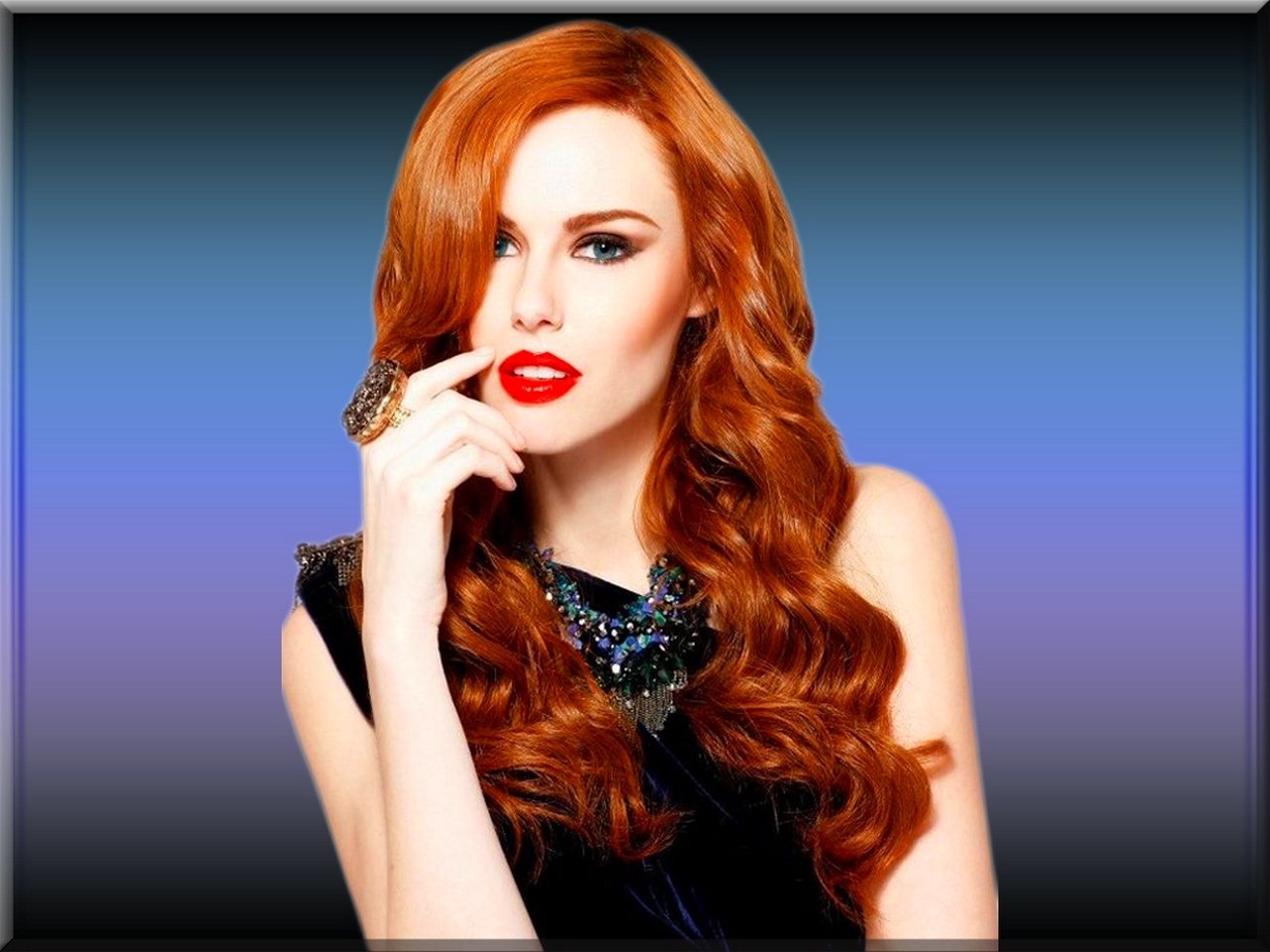 Redheaded model with long wavy hair and bold red lips, posing with a finger to her mouth against a blue-to-purple gradient background.