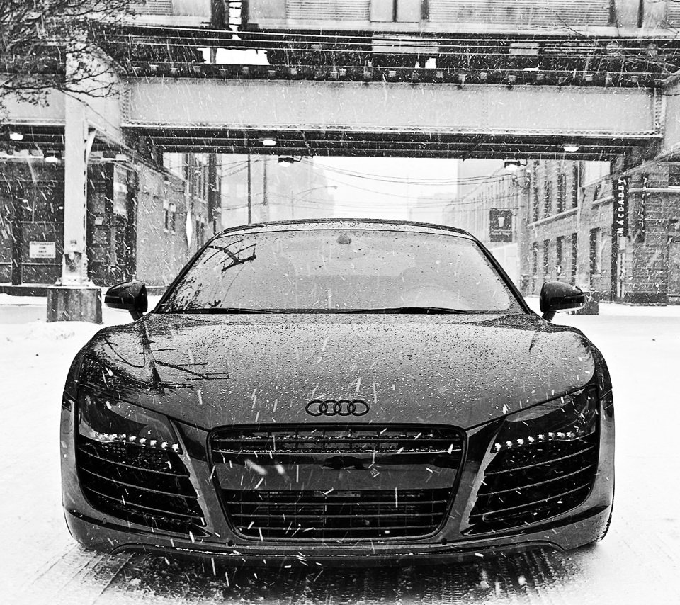 Black Audi R8 vehicle parked on a snowy urban street beneath an overpass with snowflakes falling around.