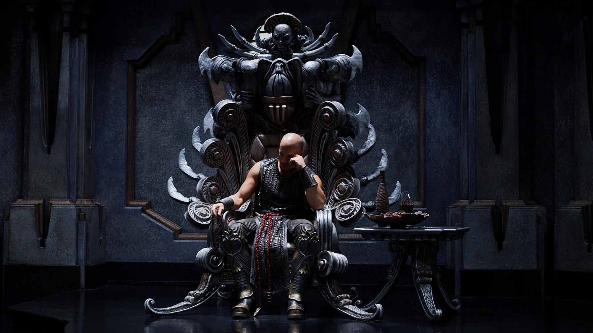 A character from the movie Riddick sits on an elaborate, dark throne in a dimly lit, ominous setting.