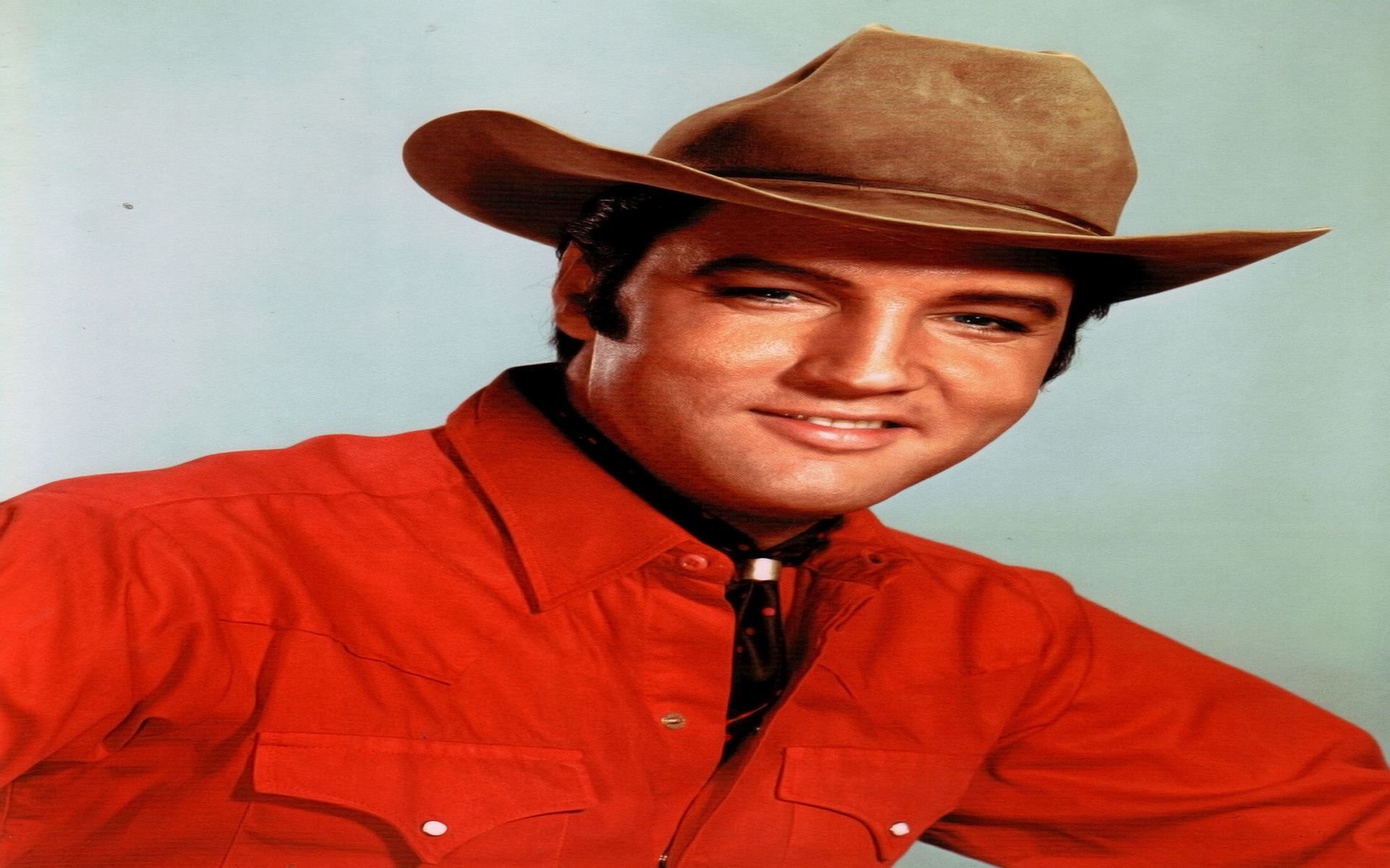Elvis Presley, the iconic Rock & Roll musician, wears a cowboy hat and red shirt, smiling confidently against a light blue background.
