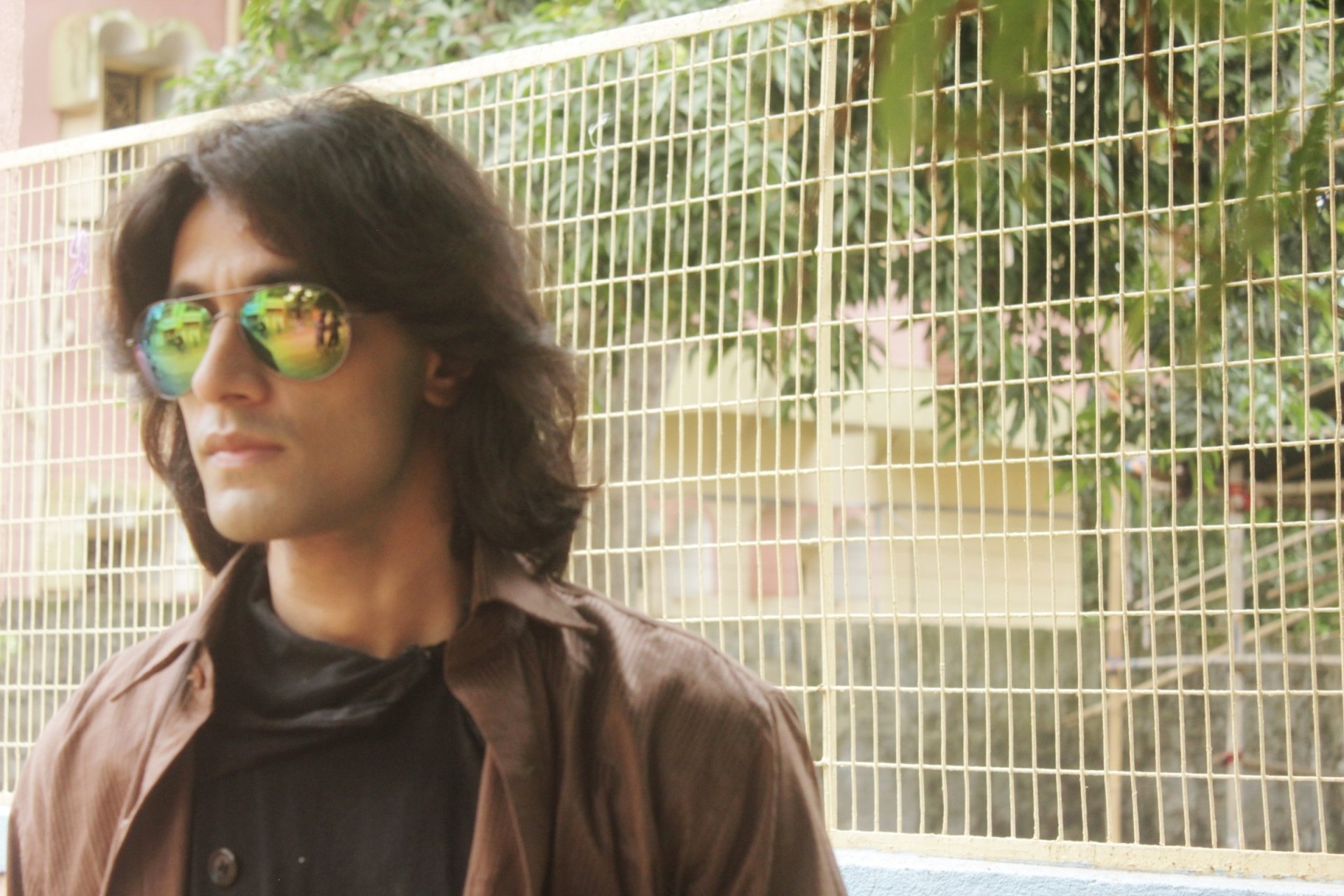 A Bengali man with long hair stands confidently, wearing sunglasses and a brown jacket, with a backdrop of greenery and a fence.
