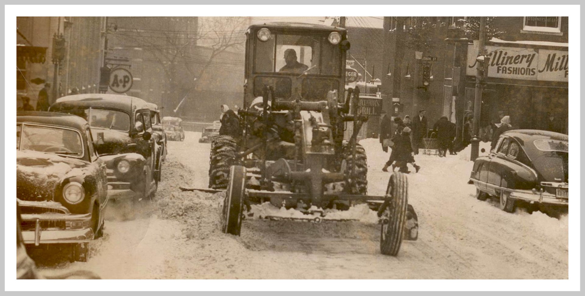 Vintage grator grader vehicle plowing snow down a city street, passing parked cars and pedestrians in a sepia-toned winter scene.