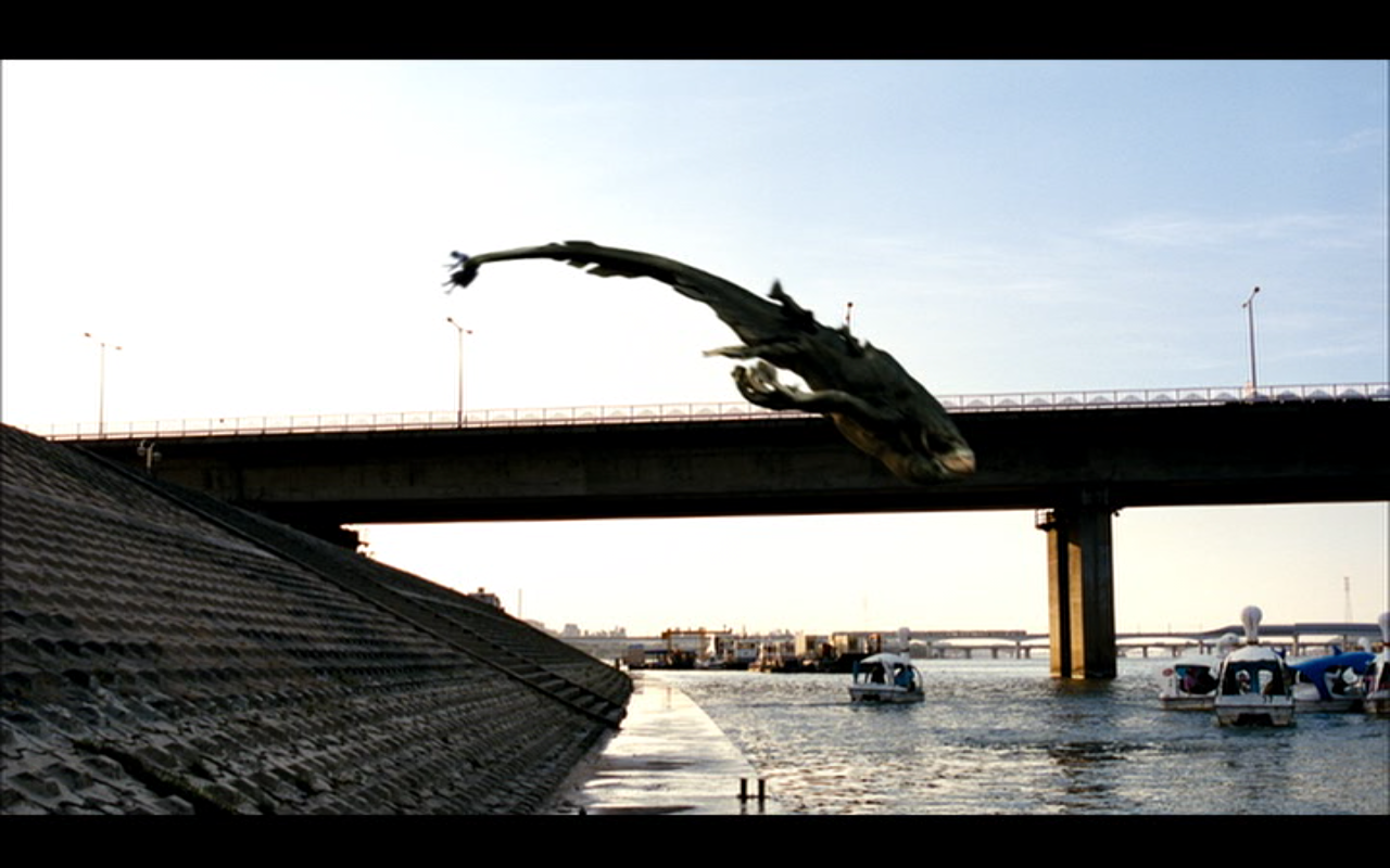 A scene from the 2006 movie The Host shows a large creature leaping from a riverside embankment toward a bridge over a river filled with boats.