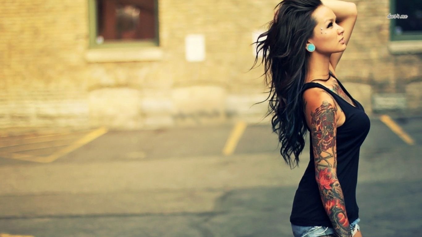 A woman with long hair poses confidently, showcasing her intricate sleeve tattoo. She wears a black tank top and earrings, against an urban backdrop.