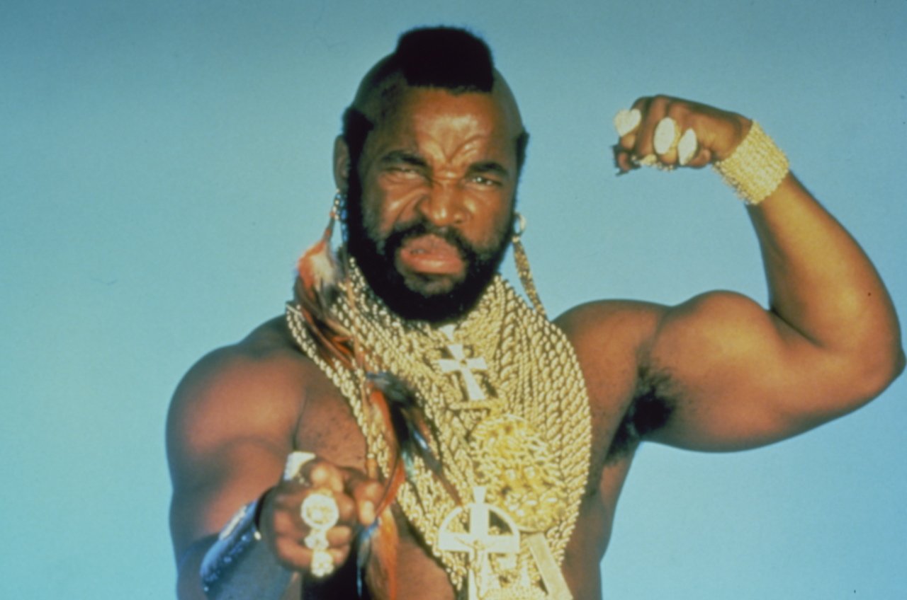 Mr. T, the celebrity, poses confidently flexing his muscles, adorned with multiple gold chains, rings, and a mohawk hairstyle against a plain blue background.