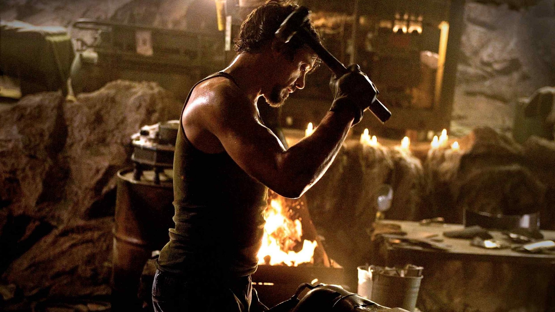 A scene from the movie Iron Man shows a man hammering metal in a dimly lit workshop, surrounded by machinery and the glow of a fire.