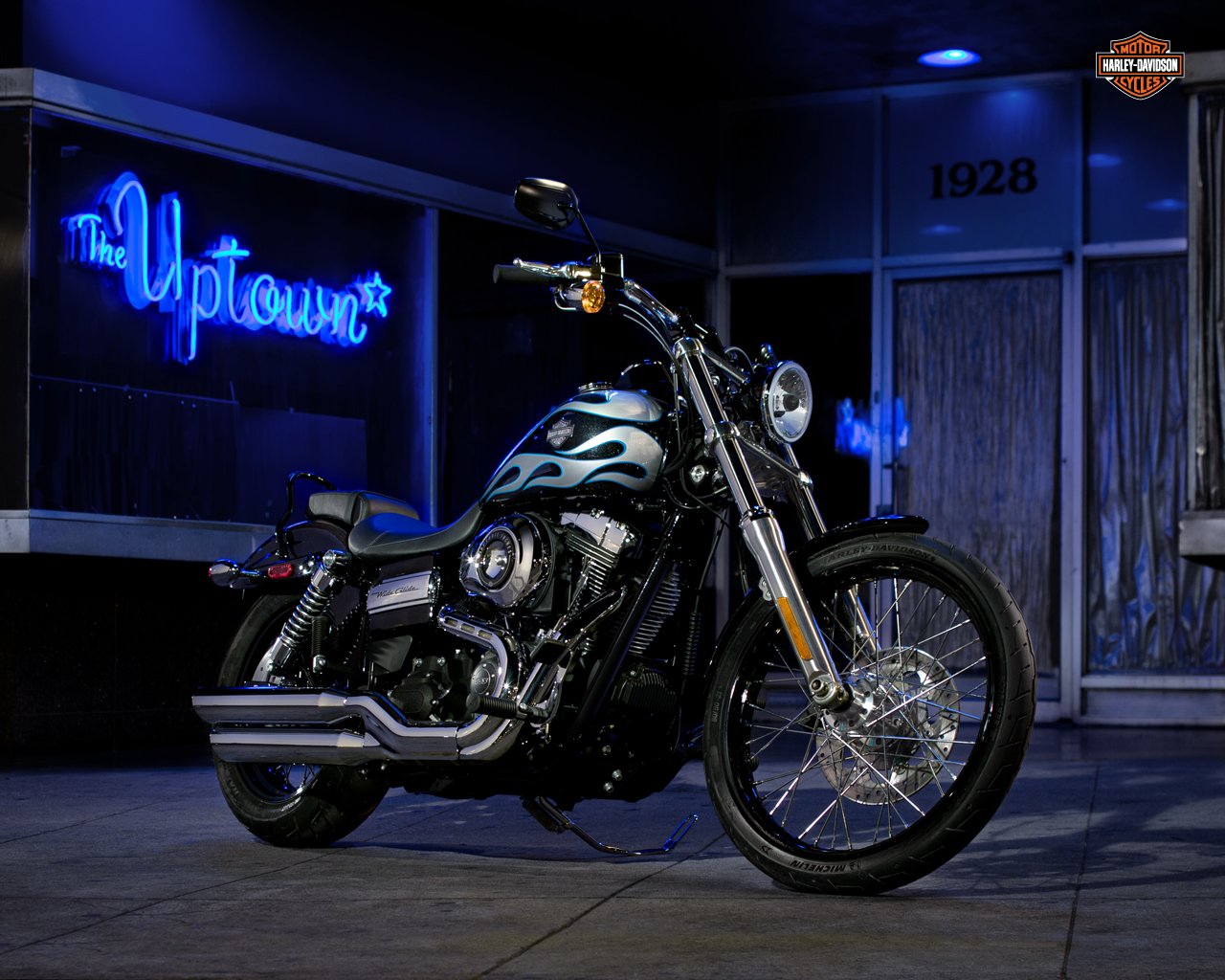 A Harley-Davidson motorcycle is parked in front of a neon sign reading The Uptown. The bike features a sleek design with blue flames, set against a nighttime urban backdrop.