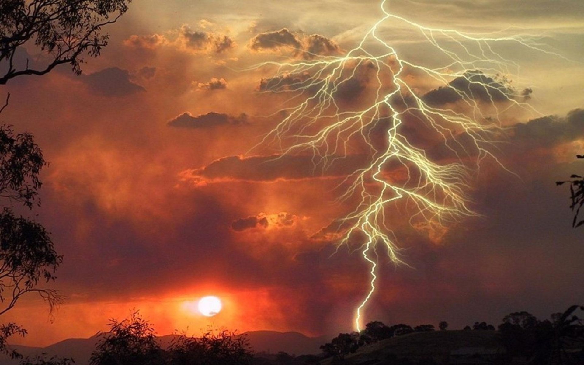 A dramatic photography capture of multiple lightning bolts striking over a silhouetted landscape at sunset with vibrant orange and red clouds.